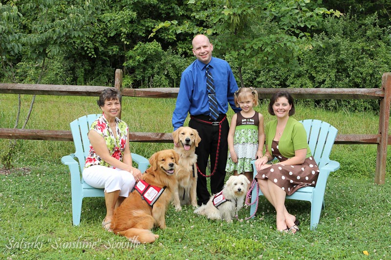 Two women seated, a man and girl standing, with two Golden Retrievers and a terrier in vest. Photo is on a grassy lawn.