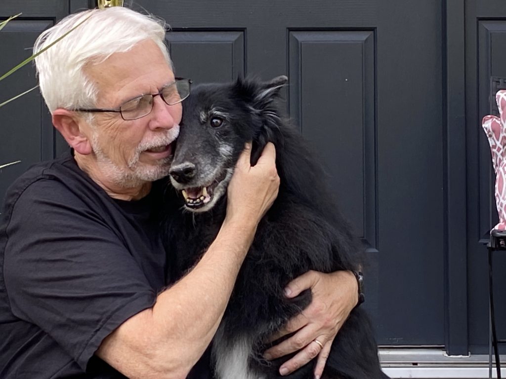 Man in a black shirt with his cheek against a black dog's cheek