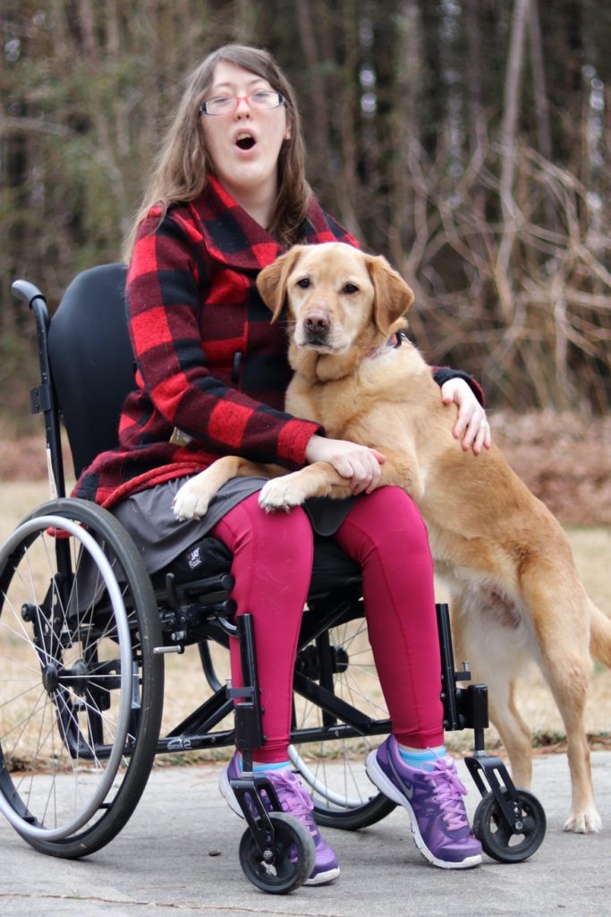 Yellow labrador with its front paws in the lap of a woman wearing a red and black plaid jacket and sitting in a wheelchair.