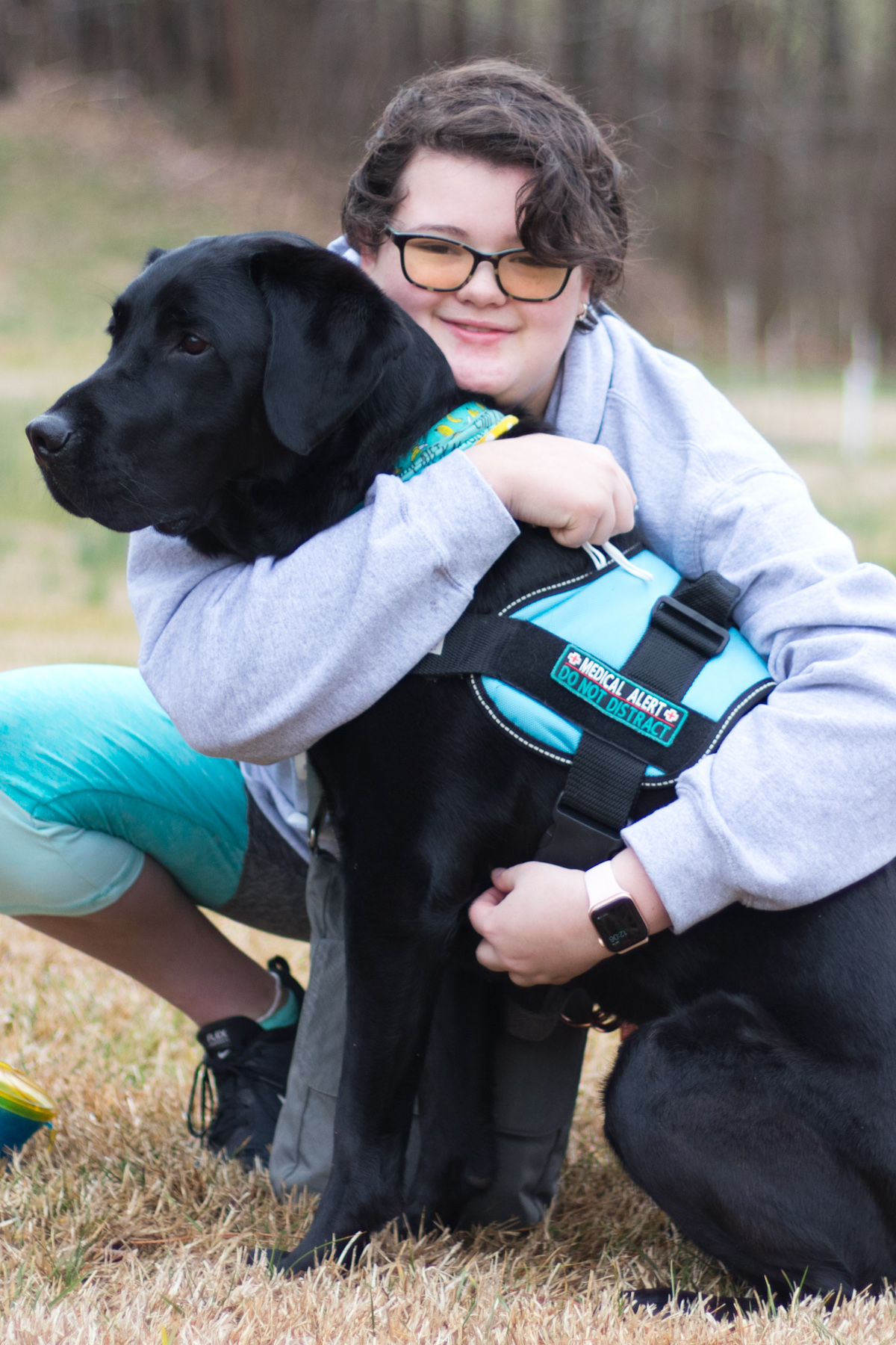 Young woman in a grey sweatshirt and teal tights with her arms wrapped around a black lab