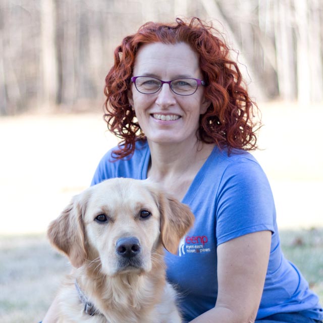 Woman with shoulder length curly red hair wearing a blue EENP shirt and purple glasses. She has her arms around a golden retriever sitting in front of her.