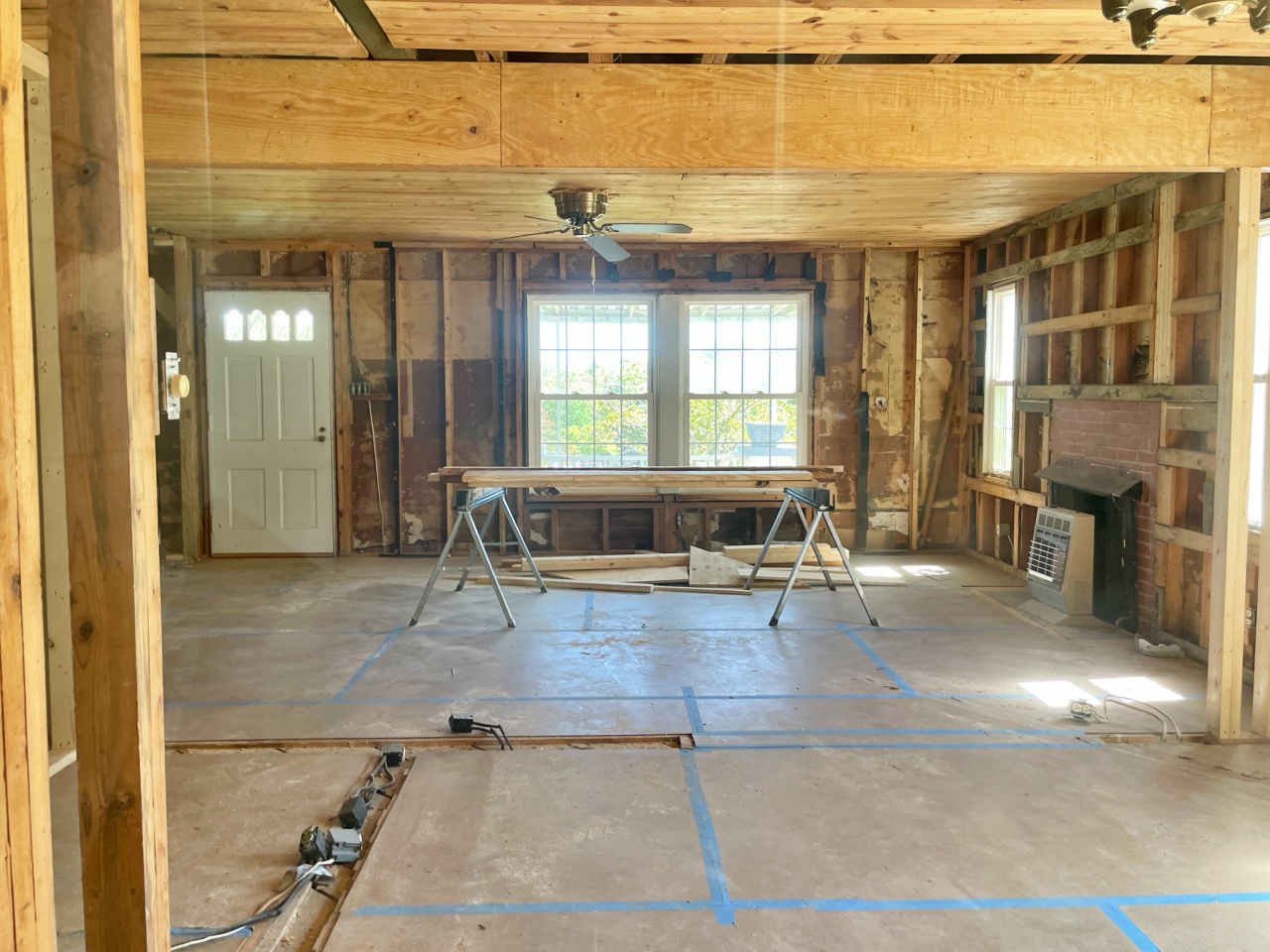 A large room under construction. The drywall has been removed, exposing the studs, and the floor is covered in protective panels. There is lumber on sawhorses in front of a double window.