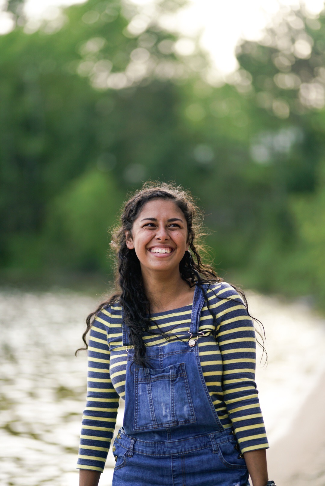 A woman with medium length brown curly hair laughs at the camera. She is standing outside with trees in the background, wearing denim overalls and a yellow and black striped shirt.