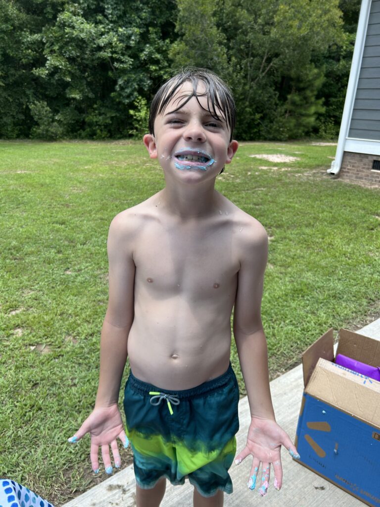Boy with brown hair standing on a concrete pad outside a house. He is wearing swim trunks and has wet hair, and his mouth and hands are smeared with blue icing. He is grinning at the camera to display the icing on his face.
