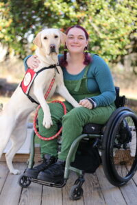 A yellow labrador retriever wearing a red vest is standing on her hind legs with her front legs in the lap of a woman with reddish purple braided pigtails and is wearing green overalls over a blue shirt. She is sitting in a manual wheelchair. A yellow labrador on the left and a woman in green overalls and a blue shirt on the right are oriented toward each other but have their faces turned to the camera. The woman is smiling with her head tipped and resting on her hand, and she is sitting in a manual wheelchair.