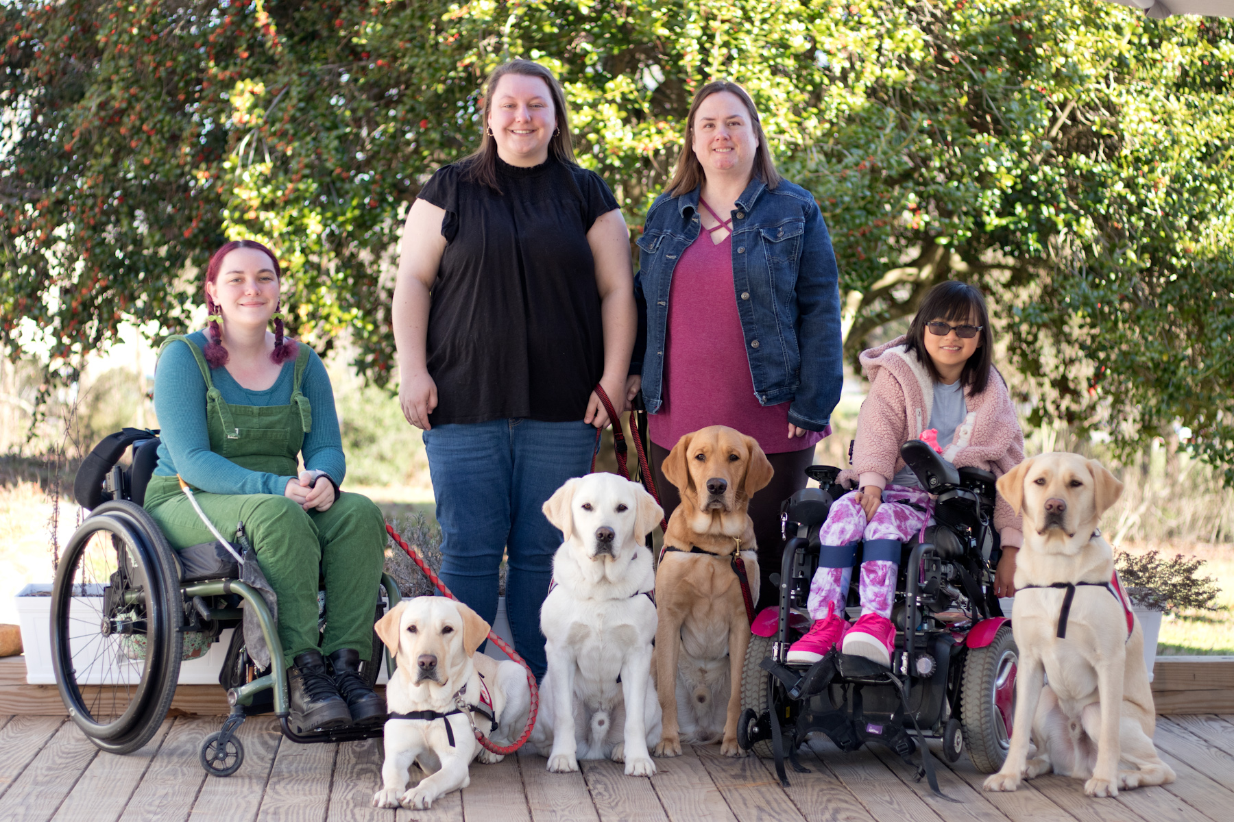 Three women and a girl posed in a row on a deck with four yellow labrador retrievers sitting in front of them. The woman on the left has reddish purple braided pigtails and is wearing green overalls over a blue shirt. She is sitting in a manual wheelchair. In the middle are two women with long brunette hair standing, the one on the left in a black blouse and the one on the right in a pink blouse with a jean jacket. The girl on the right has black hair and sunglasses and is wearing a pink jacket, patterned pink pants, and bright pink shoes which match her pink power wheelchair.