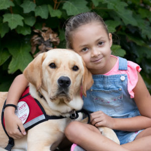 Young girl in a pink shirt and overalls with her arms around a yellow labrador dog