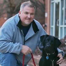 Man in a grey fleece top kneeling with his arm around a black labrador