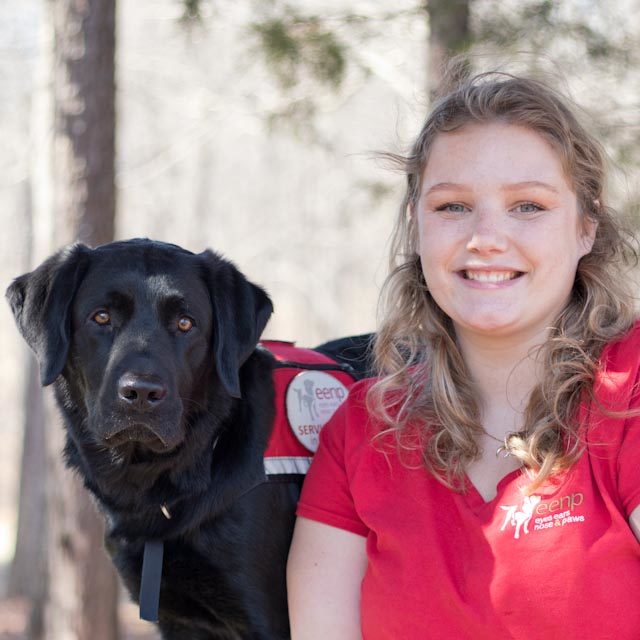 Woman with medium length brown hair in a red EENP shirt, with a black dog next to her