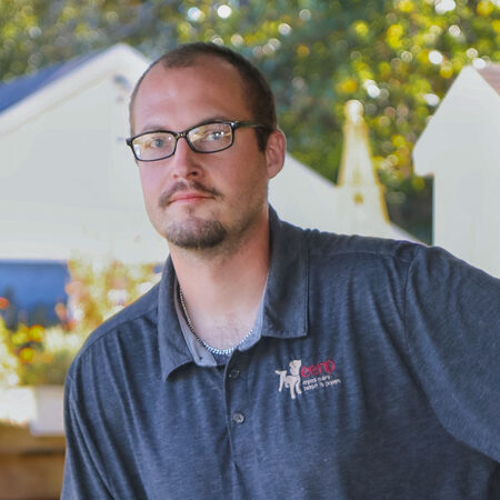 An adult man wearing glasses and a short beard is standing outdoors, leaning slightly to one side, and wearing a dark polo shirt with an EENP logo on the chest, with blurred houses and trees appearing in the background.