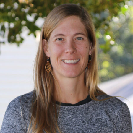 A woman with long, straight hair is facing the camera, wearing a gray sweater and leaf-shaped earrings, with blurred greenery and part of a building appearing in the background.
